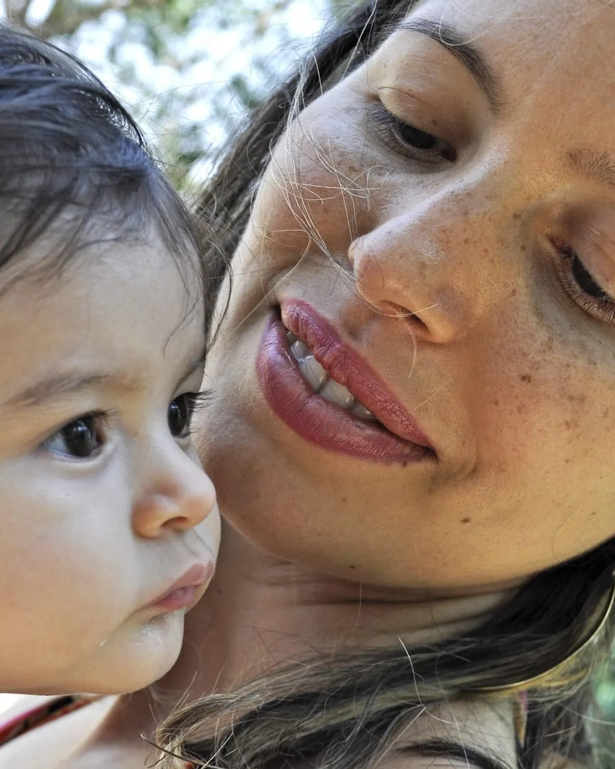 A mother holds her baby cheek-to-cheek outdoors in warm daylight, gazing down with a gentle smile.