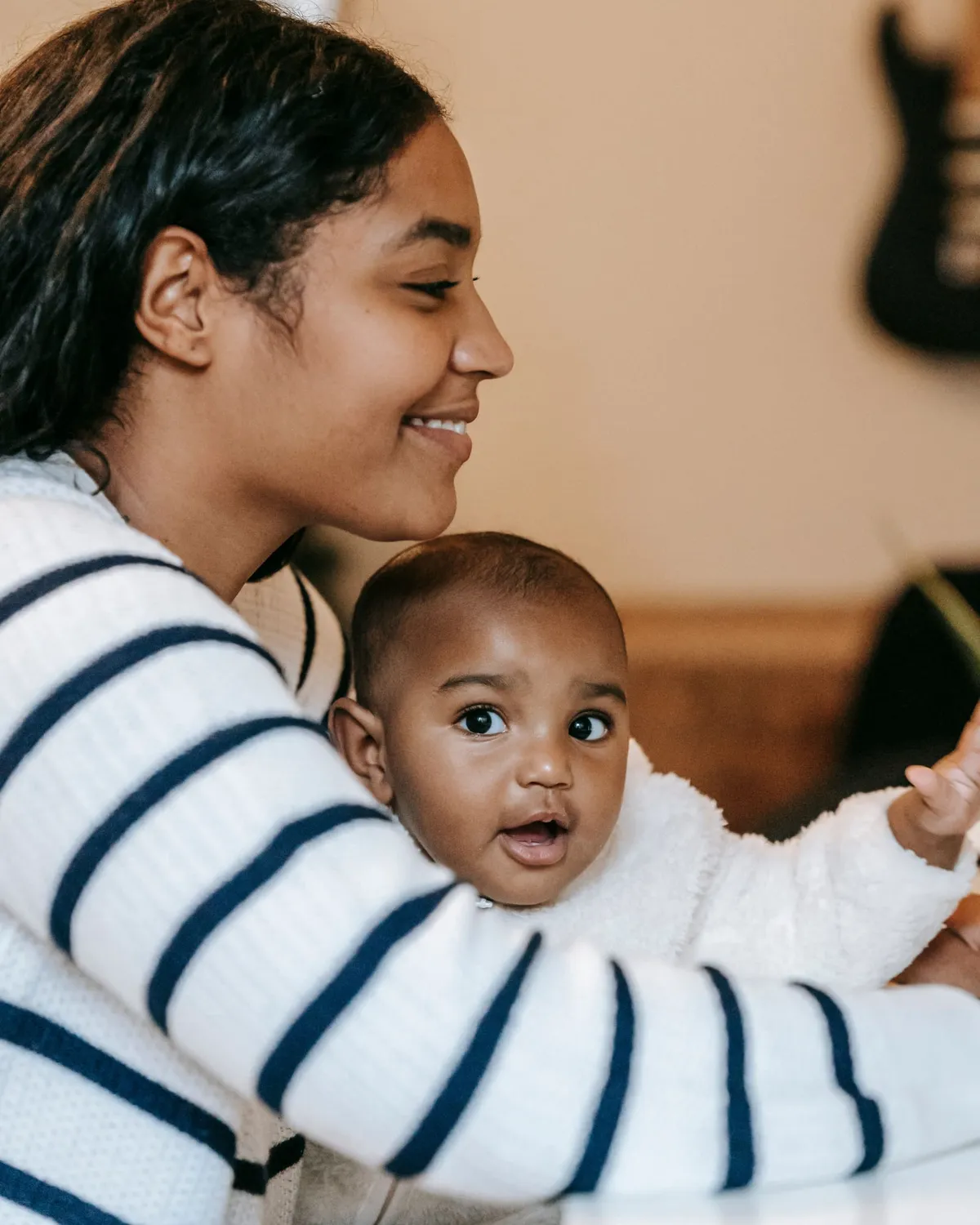 A smiling mother leans toward the baby she is holding in a warm, sunlit home as the baby reaches out.