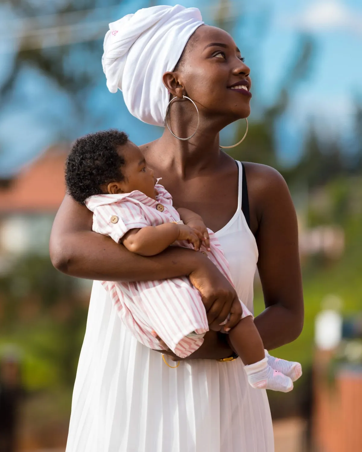 A mother in a headwrap holds her baby close outdoors in soft light, looking calmly into the distance.