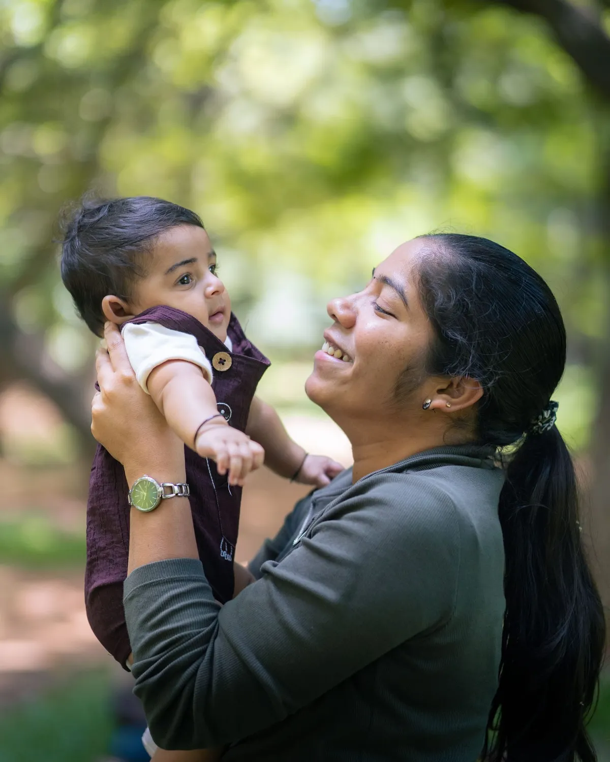 A mother lifts her baby into the air and gazes up at them, outdoors on a tree-lined path.