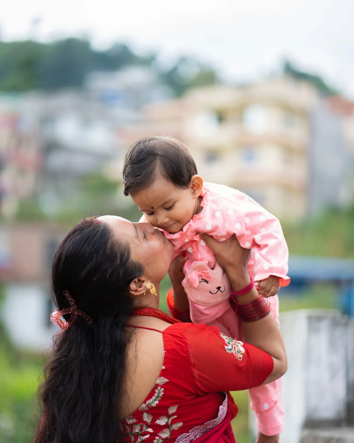 A mother lifts her baby and kisses the child's cheek outdoors, with a softly blurred hillside town behind them.